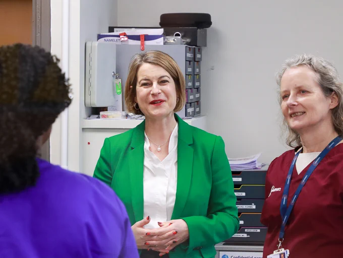 Helen Hayes MP in a green jacket, listening and speaking with two healthcare professionals inside a clinical setting.