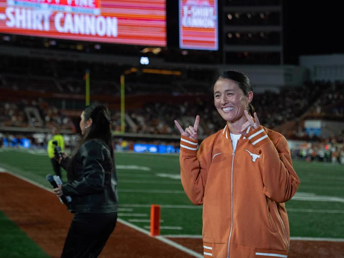 Photo of soccer coach Margueritte Bates on the DKR field at a night game. She is holding up the Hook’em sign with both hands. 

Photo by Charlie Partheymuller.