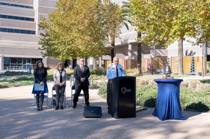 Four individuals stand outdoors at a podium labeled "San Bernardino County," with one person speaking into the microphone. They are surrounded by trees and buildings, in a well-lit, landscaped area. A table draped with a blue cloth is positioned to the right.