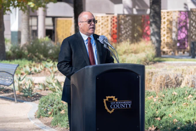 Person speaking at a podium with the San Bernardino County logo, outdoors during daytime.