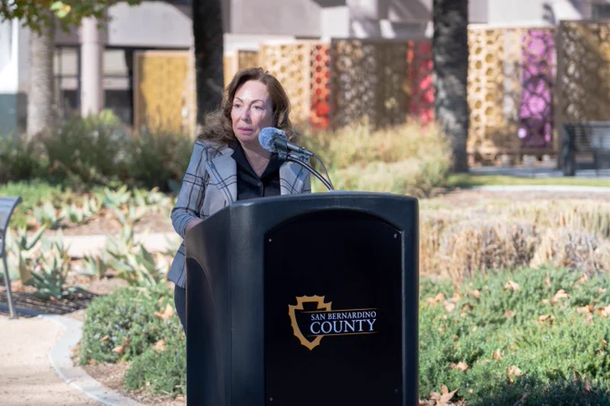 A person speaking at a podium branded with the "San Bernardino County" logo, set in an outdoor setting with plants and buildings in the background.
