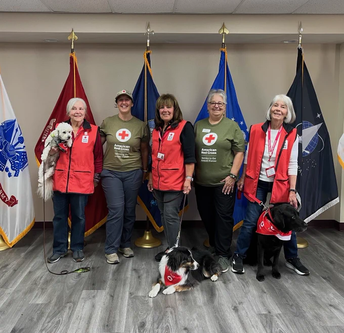 Five American Red Cross volunteers, accompanied by three service dogs wearing Red Cross vests, standing in front of a row of various military flags.
