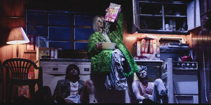 Image showing three individuals in a dimly lit room sitting around a small table covered in snacks, including a box of lucky charms. One person is wearing a fuzzy green outfit, one is in casual attire, and another is wearing a mask, creating a unique, moody atmosphere.