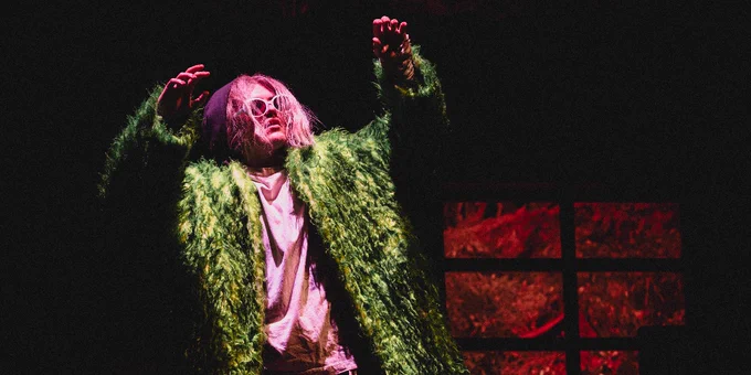 Person in a green fur coat and sunglasses performing dramatically on stage with red lighting in the background.