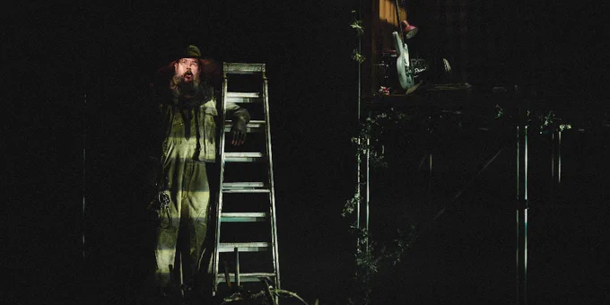 Scene from a theatrical production featuring a person in overalls standing next to a ladder on a dimly lit stage, with a second person partially visible in the background.