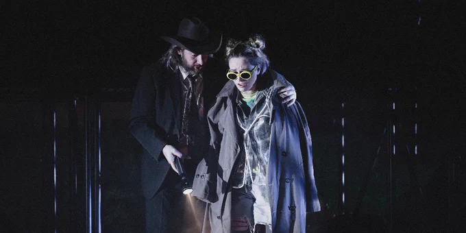 Two people on stage in a theatrical production, one wearing a hat and a coat, assisting the other who wears sunglasses and a decorative scarf, illuminated by dramatic stage lighting.