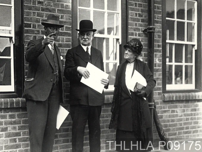 Black and white photograph of George Lansbury (centre), social reformer and politician, and his wife Bessie attending the opening of Summer House in Maddams Street, Bromley. Both Lansburys are clutching pieces of paper and are listening to an explanation from a third unidentified individual in the picture (ref. P09175).