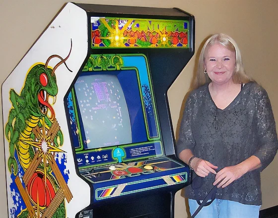 Blonde woman smiling next to a vintage arcade cabinet with colorful insect artwork and a glowing game screen.