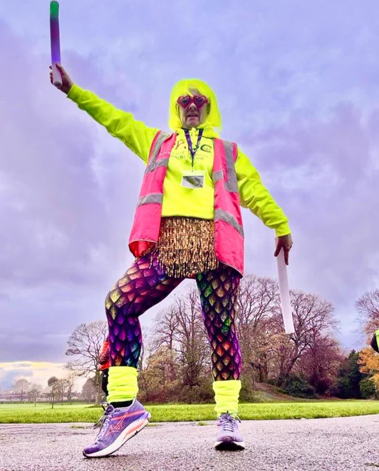 A parkrun volunteer posing in colourful attire including a neon yellow wig and bright leggings with her right arm up.