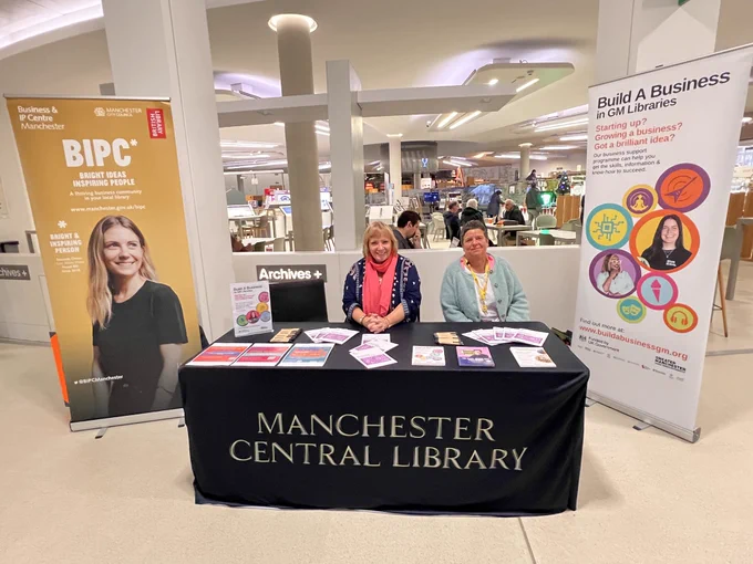 2 members of Business team sat at stall promoting business support with 2 pop up banners on either side of stall. Table cloth states Manchester Central Library.