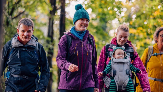 Group of five individuals, including one carrying a baby in a front carrier, smiling and walking in a colorful autumn forest.