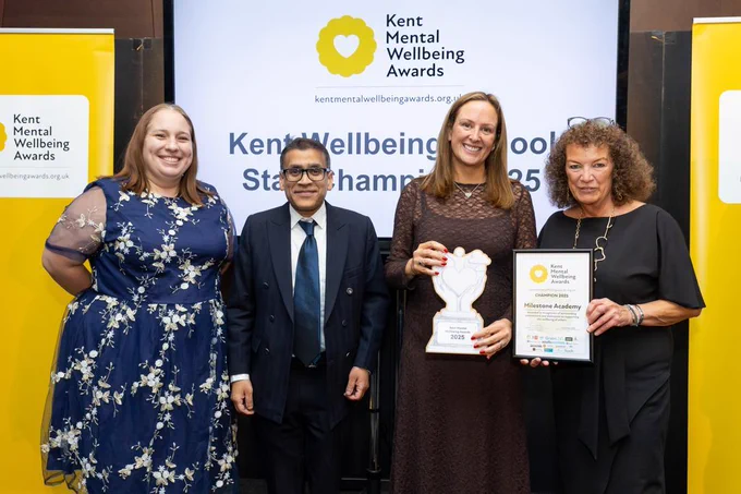 Four people smiling at the Kent Mental Wellbeing Awards, with two holding a Champion 2025 trophy and certificate in front of a Kent Mental Wellbeing Awards branded backdrop.