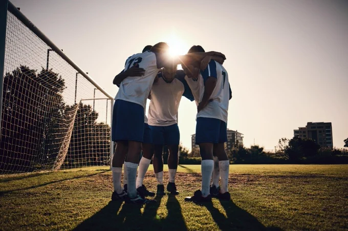 ​A warm, golden-hour shot of a grassroots football team in a huddle on the pitch. The players have their arms around each other in a circle, symbolizing unity, support, and the tangible sense of community described in the post.