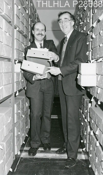Black and white photo of two men standing between archive shelves holding boxes.