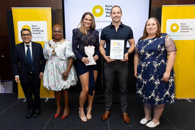 Five people smiling at the Kent Mental Wellbeing Awards, with two holding a Champion 2025 trophy and certificate in front of a branded backdrop.