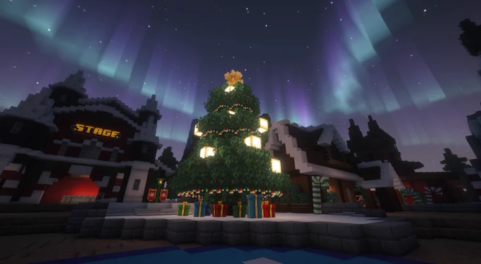 A large Minecraft Christmas tree decorated with glowing lights and presents underneath, set in a snowy village square at night with colorful aurora lights in the sky.
