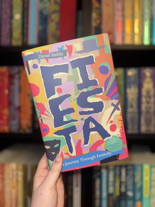 A hand holding the book Fiesta: A Journey Through Festivity by Daniel Stables in front of a blurred bookshelf. The cover is colourful and abstract, with bold painted letters spelling “FIESTA” against a vibrant background of splashes and shapes.