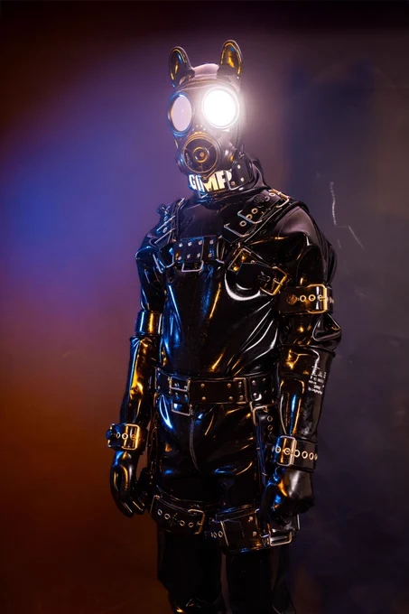 GimpSimp in full black heavy rubber, wearing bondage harnesses cuffs and a gasmask. One lens reflects shines at the camera