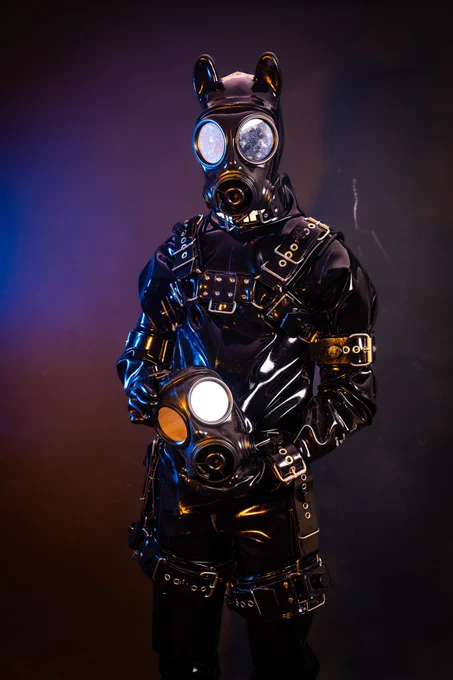 GimpSimp in full black heavy rubber, wearing bondage harnesses cuffs and a gasmask. He holds another gasmask