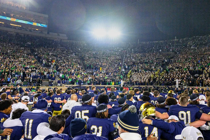 Football players from the Notre Dame team gather on the field with their backs to the camera, facing a large, excited crowd in the stadium stands at night. Both the players and the crowd have their arms around one another as they sing the Alma Mater. 
