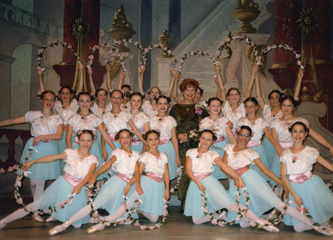 Group of young ballerinas in pastel tutus posing with floral hoops on stage in front of a painted backdrop.