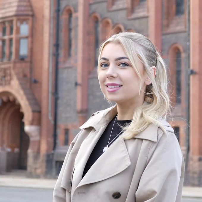  A photograph of article author, Millie Green. Millie is stood outside a university building wearing a cream rain mac and black top. She has blonde hair which is up in a ponytail.
