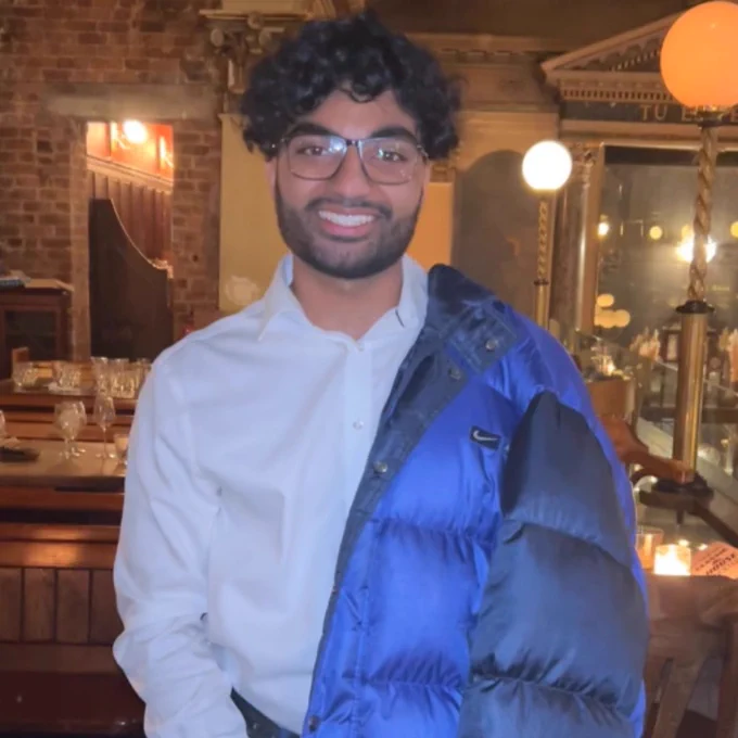 A photograph of article author, Sam Dhir. Sam is stood up wearing a white shirt and jeans, with a blue puffer jacket draped over one shoulder. He has dark curly hair and glasses.