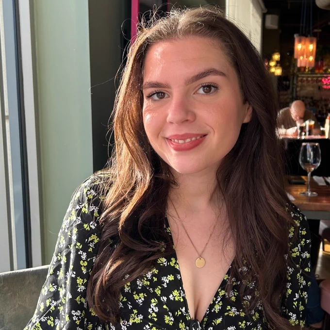 A photo of article author, Lily Robson. Lily is sat by a window wearing a dress with a floral pattern and a gold necklace. She has long brown hair and is smiling at the camera.