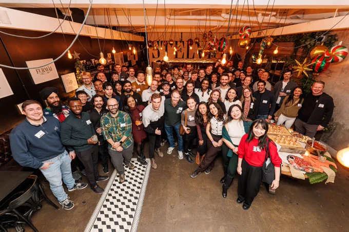 A large group of people smiling at a YC founders’ holiday party in Toronto, gathered in a warmly lit venue with festive decorations and food in the background.