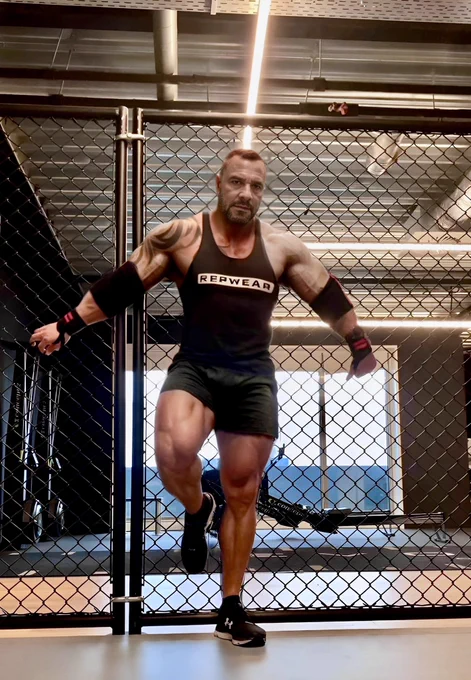 a muscular man standing in front of a chain-link cage in a dimly lit gym. His pose is confident and open, with strong body language suggesting intensity and presence. The dramatic lighting and setting create a bold, inviting atmosphere.