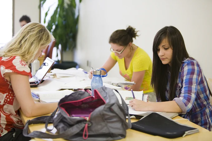 Three students studying and taking notes at a table surrounded by books and backpacks.
