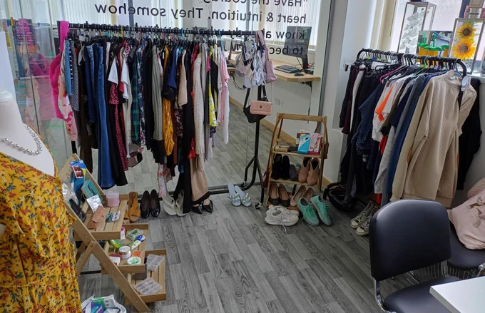 Clothing racks filled with various garments and shoes displayed in a bright boutique with wooden flooring and a mannequin in a yellow floral dress.