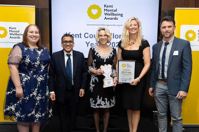 Five people are stood in front of a Kent Mental Wellbeing Awards banner, with the two winners holding up a wooden trophy and a winner's certifcate