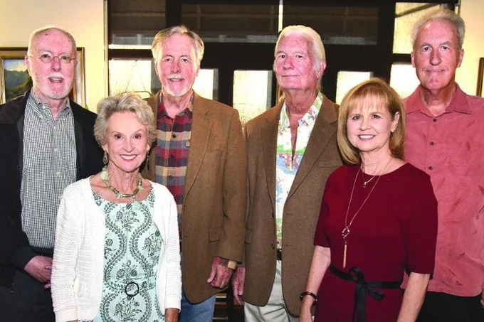 Group of six adults smiling for a photo in an indoor setting, standing in a line with various styles of casual and semi-formal attire.
