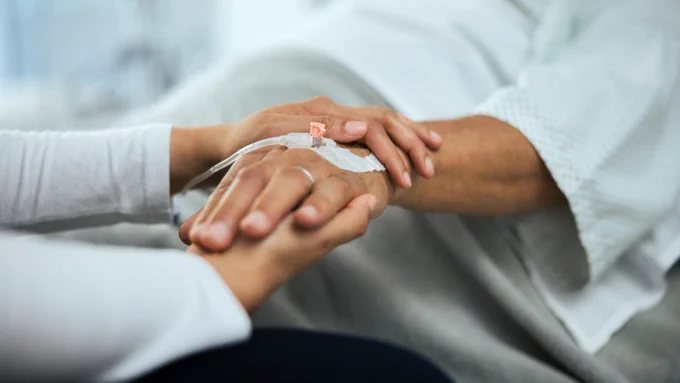 Close-up of a patient’s hand with an IV line being gently held by a caregiver in a hospital setting.