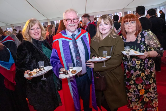 Image shows a PhD graduate in his red, blue and purple gown, accompanied by his family. They are all holding plates and some are holding glasses of bubbles.