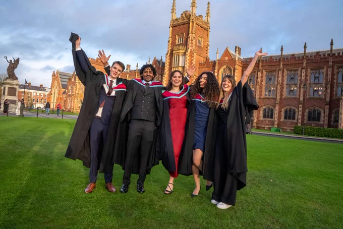 Image shows a group of graduates jumping for joy on the lawn at the front of the Lanyon Building. There are two men and three women and they are all wearing their graduation gowns.