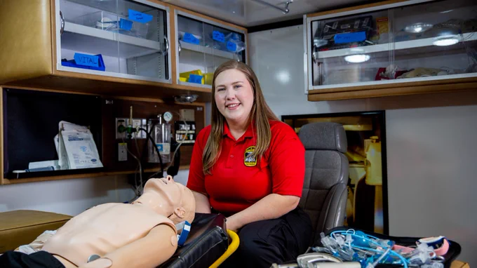 Person in a red polo shirt seated next to a medical training mannequin in an ambulance, surrounded by medical supplies.