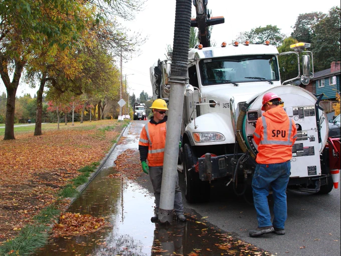 Two utility workers from Seattle Public Utilities next to a truck equipped with machinery, cleaning a leaf-filled street gutter in a residential area with autumn foliage.