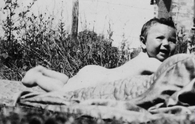 A vintage black and white photo of a young child reclining on a blanket outdoors, smiling at the camera.