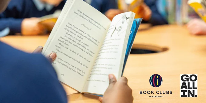 A close up of a child's hands holding an open book. There are other children reading in the background. 'Go All In' logo for the National Year of Reading and the Book Clubs in Schools logo. 