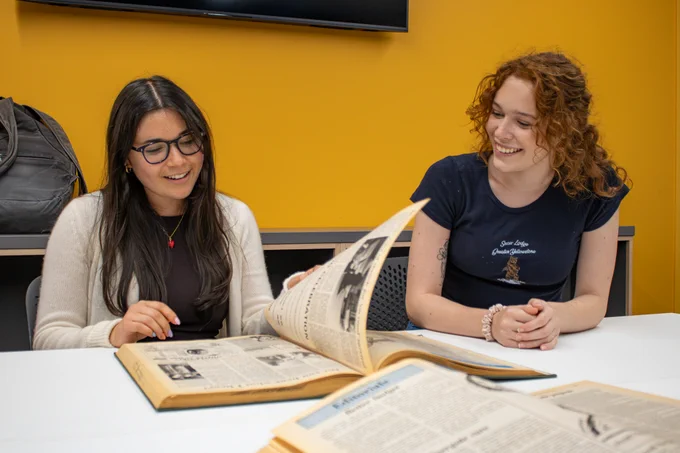 Two people sit at a table flipping pages of a bound newspaper