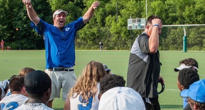 A group of young players sit on the edge of a football field while two coaches speak to them. One coach raises his arms in encouragement, the other leans forward as he talks. It's a candid moment of community sport, showing motivation, teamwork and the human side of training.