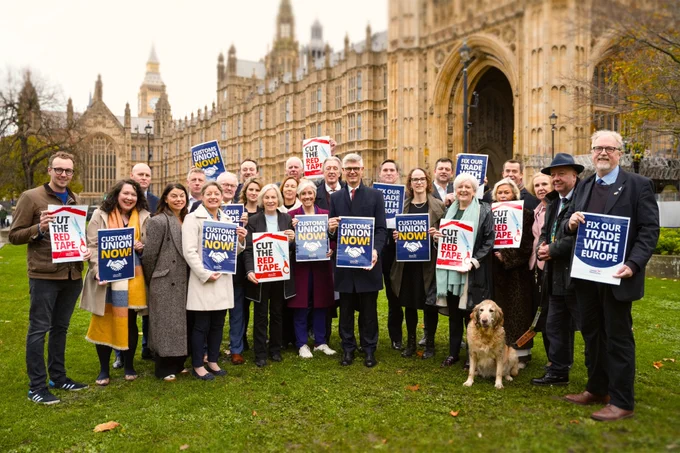Lib Dem MPs stand on the grass outside the Houses of Parliament holding various red, white, and blue signs that say "CUSTOMS UNION NOW!," "CUT THE RED TAPE," and "FIX OUR TRADE WITH EUROPE."