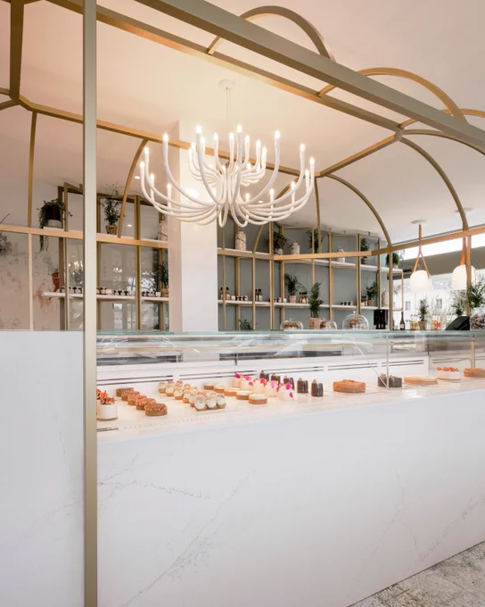 A brightly lit bakery interior with a white marble counter displaying various pastries and cakes. Shelves in the background hold plants and bottled goods, illuminated by a modern chandelier.