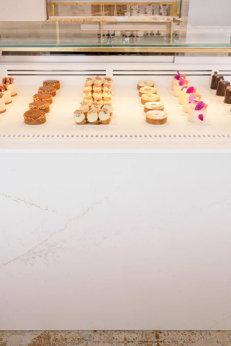 A display case presents rows of artfully arranged pastries: caramel nut tarts, cream puff clusters, glazed tarts, and petit cakes with floral accents. The lower part shows white marble and decorative tile.