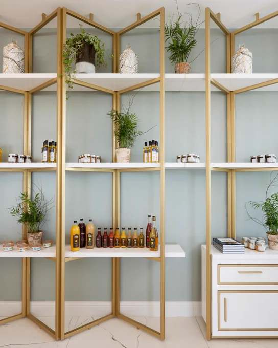 A tall gold shelving unit displays decorative items and food products on white shelves. Potted plants, jars, and bottles are arranged attractively. A white cabinet sits to the right, with stacked books on top.
