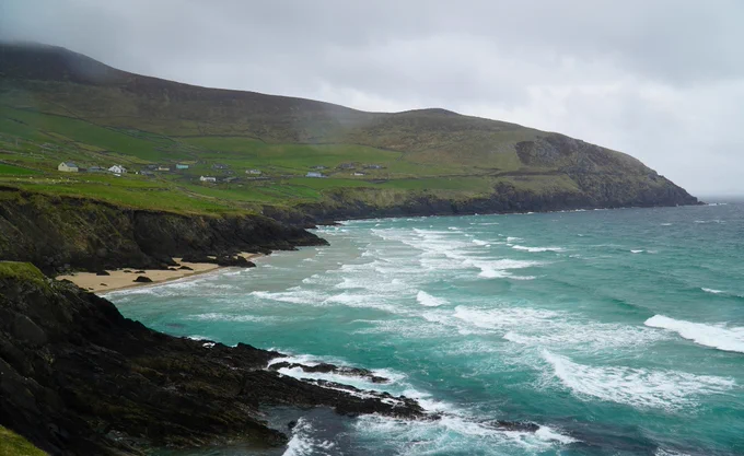 Coastal scene with a rugged shoreline and small sandy beach. Turquoise waves crash against the rocky cliffs. Green fields and scattered houses stretch across the gentle hills under a cloudy sky.