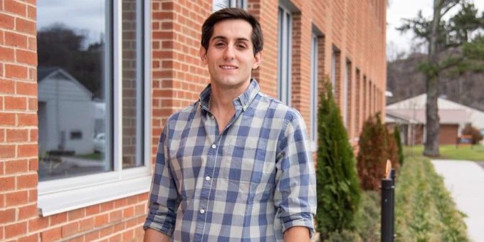 Person in a plaid shirt standing outside a building with windows and a green landscape in the background.
