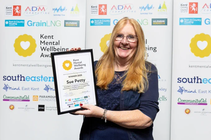 Sue is smiling and holding up her winner's certificate infront of a Kent Mental Wellbeing Awards backdrop
